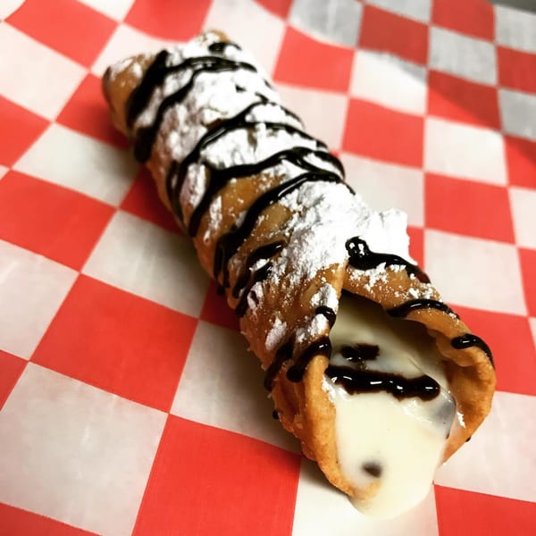 A cannoli from Jack's Pizza, drizzled with chocolate and powdered sugar, on a red and white checkered paper.