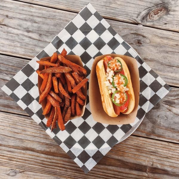 A hot dog with jalapeños and onions, alongside sweet potato fries, on a checkered paper.